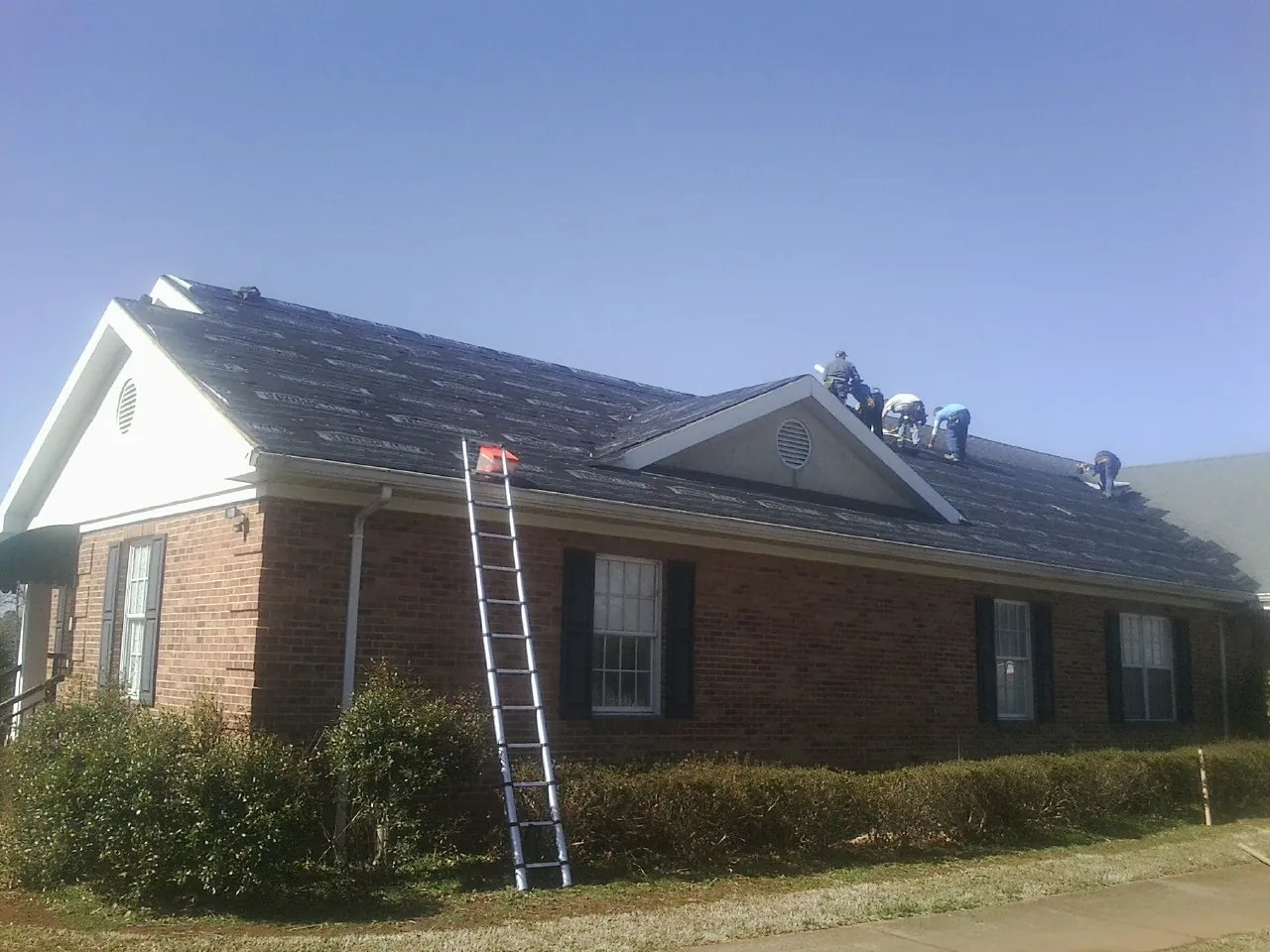 Crew tearing shingles off a brick ranch roof with a ladder set — Metal Roof Repair work by our crew in Britt