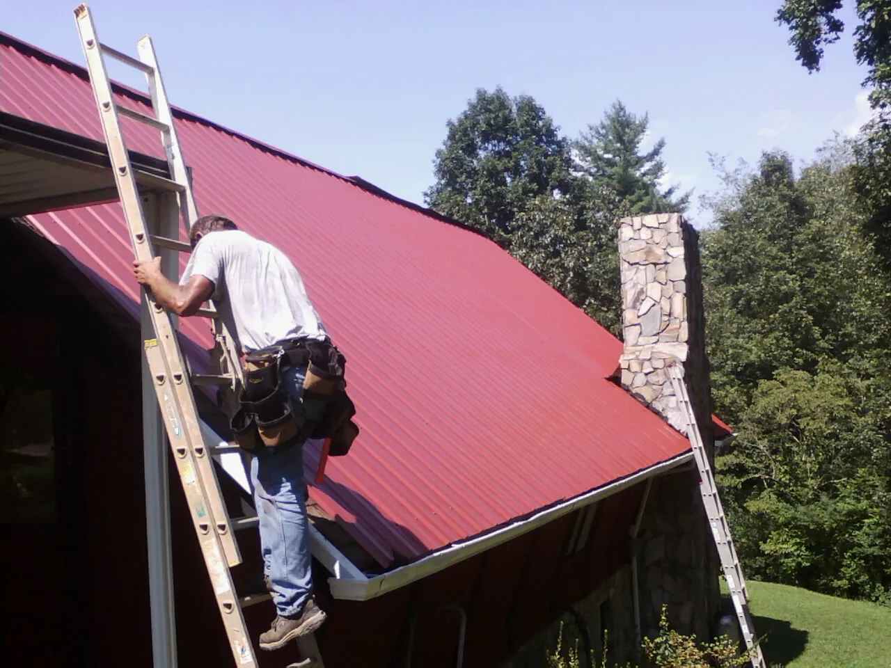 Roofer on a ladder beside a red metal roof and stone chimney — Soffit & Fascia Repair by Roofing Brothers in Britt